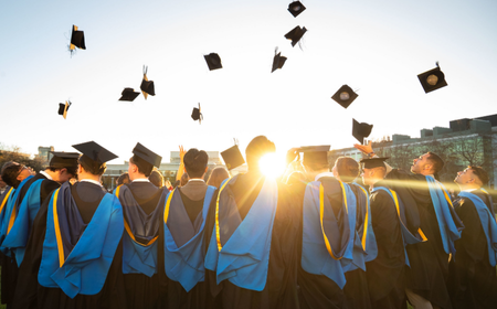 UCD alumni celebrating their graduation, standing in gowns and throwing caps into the air at sunset.